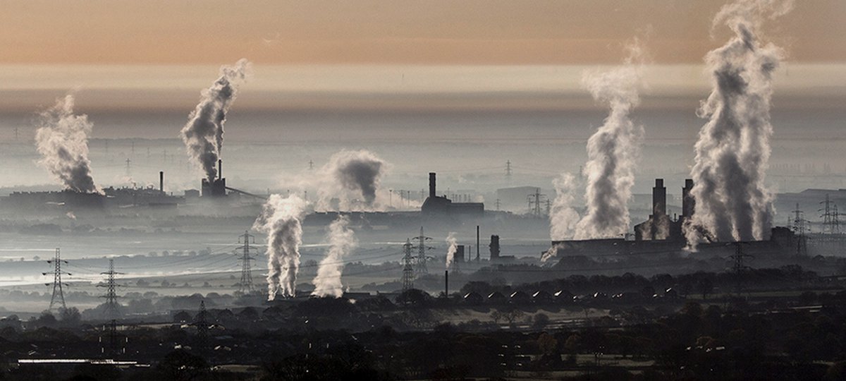 Plumes of steam rising from industrial chimneys across a hazy plain, crossed by lines of electricity pylons