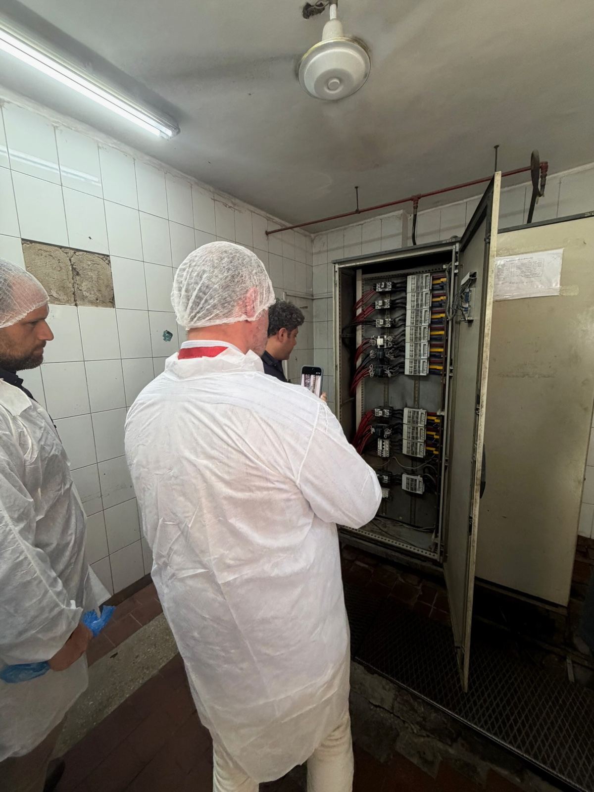 People in hairnets and white coats at an open distribution board in a dairy’s plant room, seen from behind and in profile