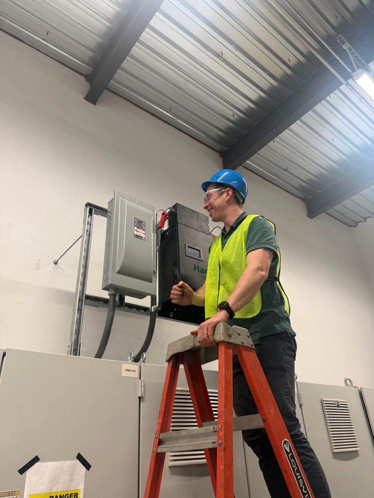 One of the HarmoniQ team in a hard hat and high-visibility vest on a stepladder, beside a wall-mounted Delta and its isolator