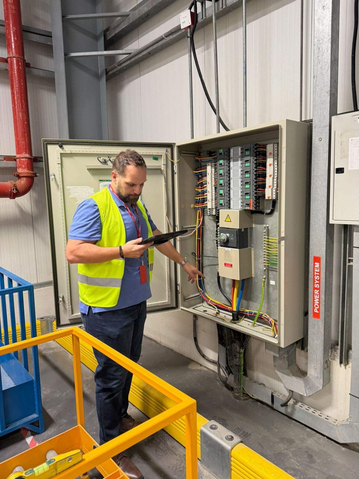 A member of the HarmoniQ team in a high-visibility vest checking an open distribution board against a tablet