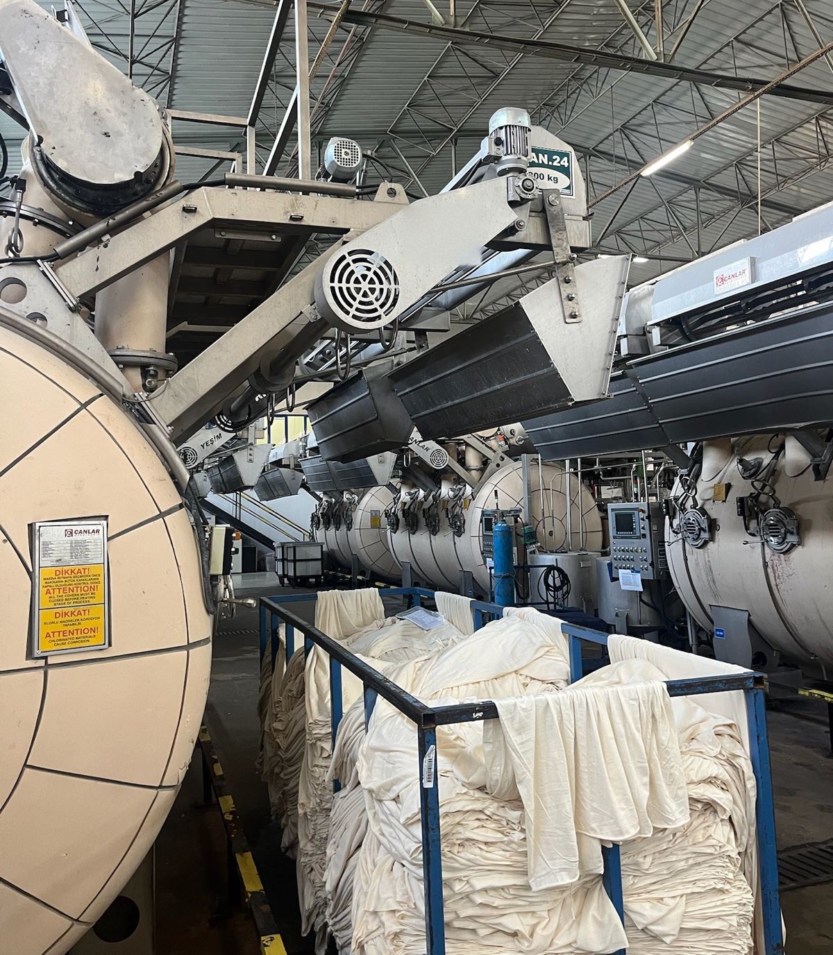 A large fabric dyeing machine in a textile mill, bins of white fabric beneath it
