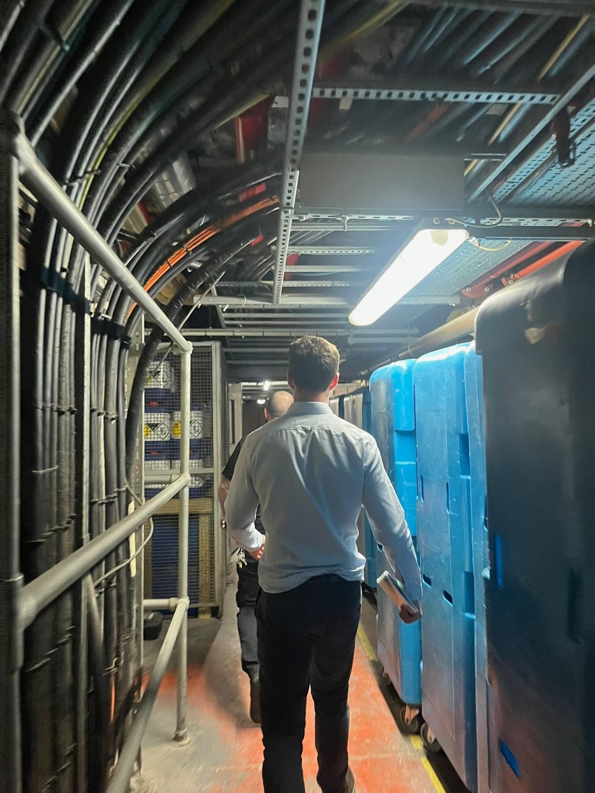 A member of the HarmoniQ team, seen from behind, walking a hotel’s basement service corridor beneath heavy cable runs
