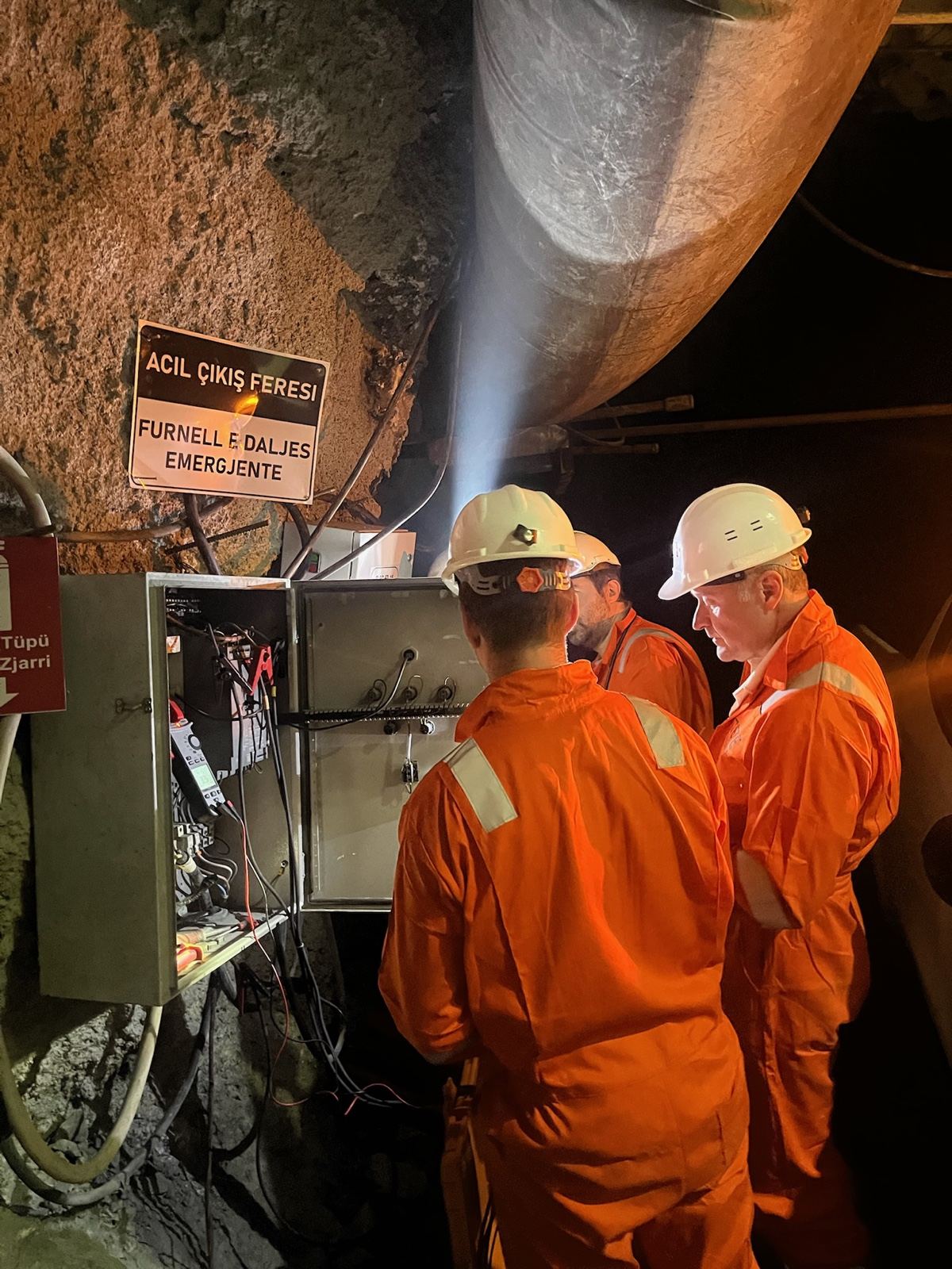 The team in coveralls and hard hats at an open panel underground, an emergency-exit sign above