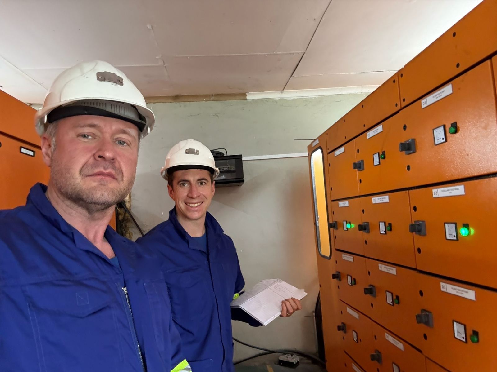Two of the HarmoniQ team in hard hats and coveralls beside a run of orange motor control panels
