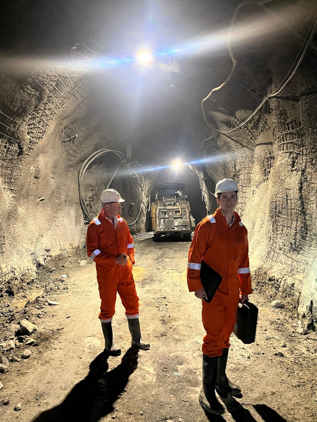 Two of the HarmoniQ team in coveralls and hard hats in a mine tunnel, a loader behind them