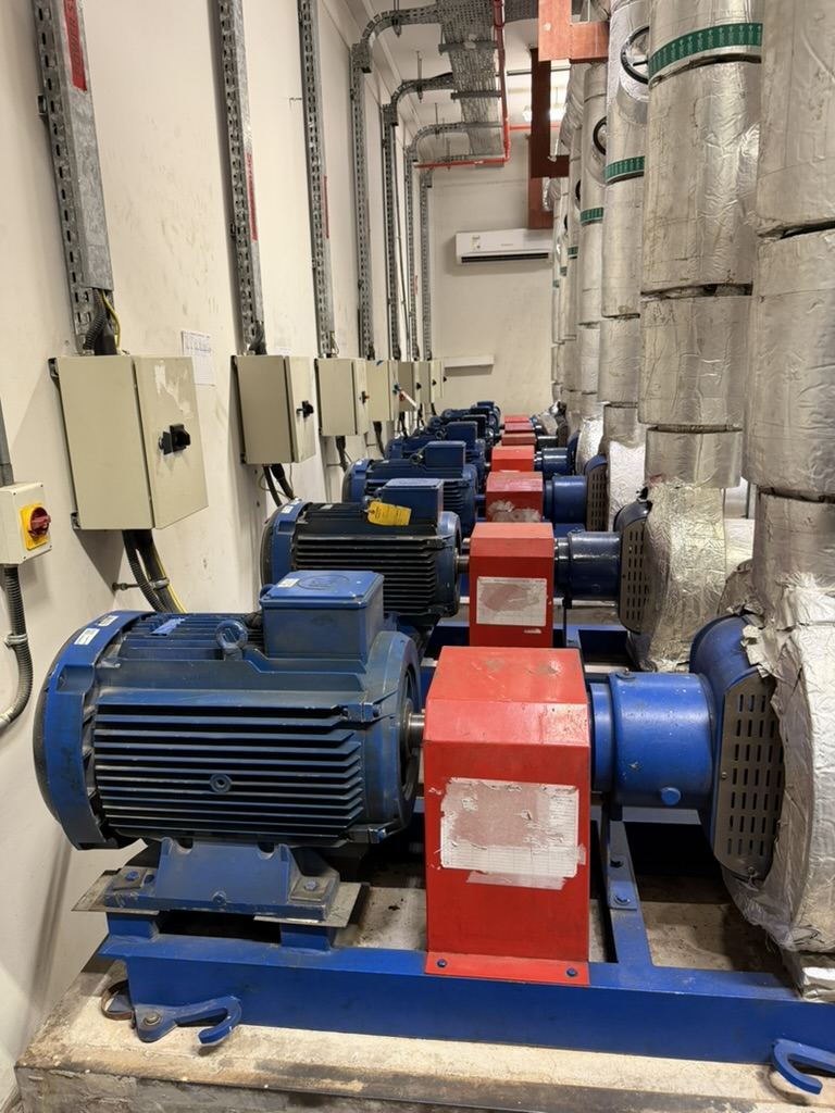 A row of blue electric motors driving pumps along a plant-room wall, insulated pipework alongside
